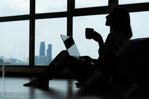 Siluet girl working on a laptop and drinking coffee, sitting on the floor near the bed by the panoramic window with a beautiful view from the high floor. A cozy workplace. Shopping on the Internet.