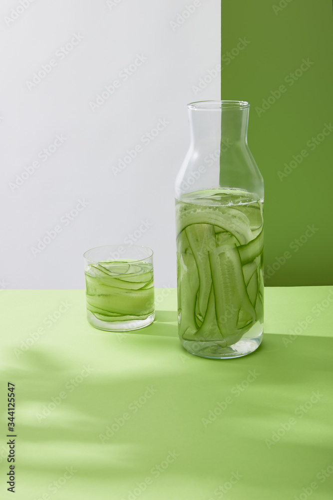 Bottle and glass with fresh drink made of sliced cucumbers on grey and green background