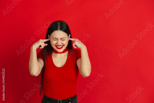 Crazy covering ears with hands. Caucasian young woman's monochrome portrait isolated on red studio background. Beautiful female brunette model. Concept of human emotions, facial expression, sales, ad. © master1305