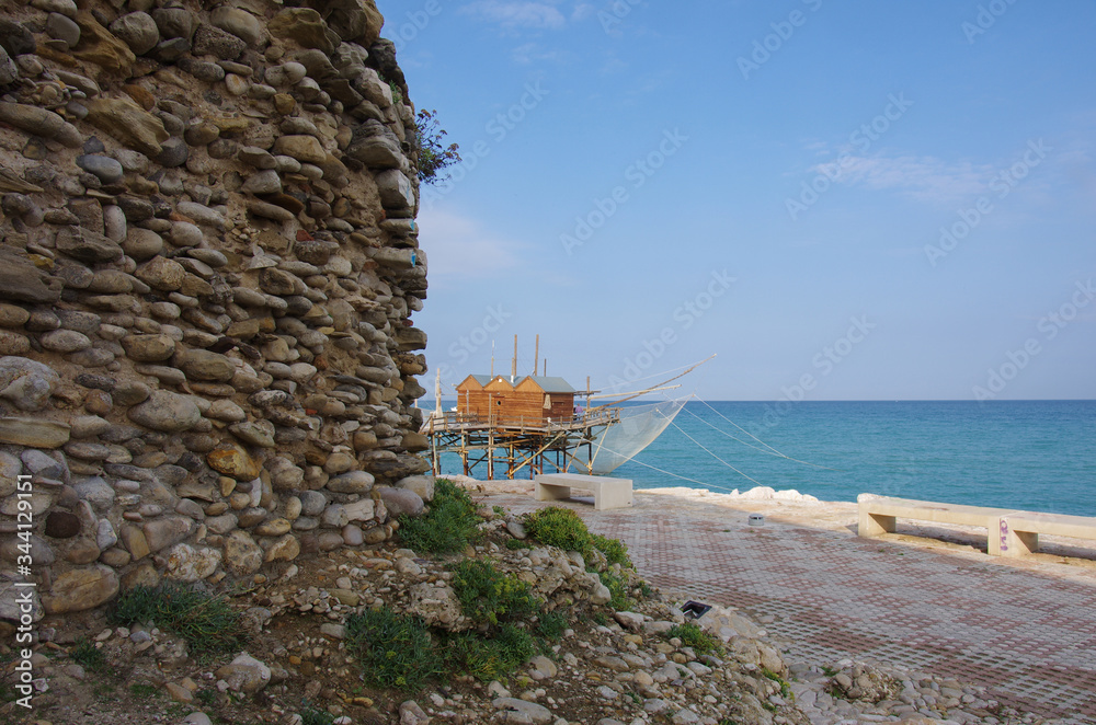In the foreground ancient stone walls and in the background a characteristic fishing trap (trabucco) of the Molise coast.Termoli, Italy.