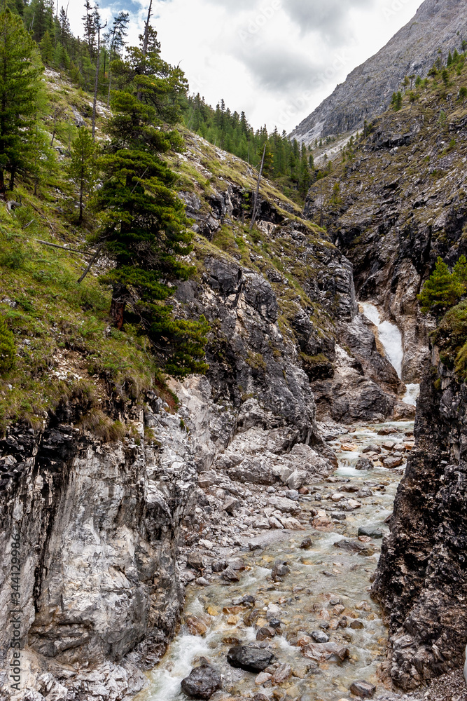 Gorge with a river in the mountains. A small waterfall at the end. Coniferous trees on the slopes. The weather is cloudy. Vertical.