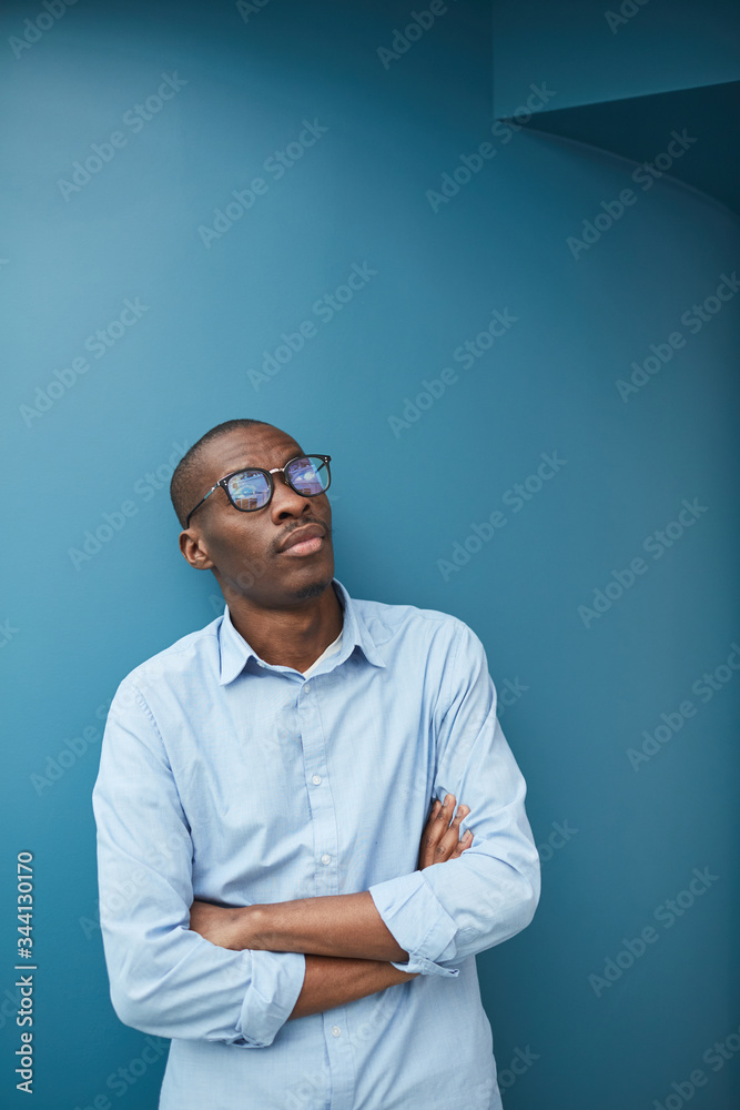 Young African businessman in eyeglasses standing with his arms crossed with pensive look against the blue background