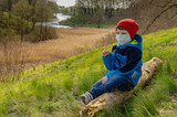 cute boy in a medical mask sits on a hill on a log and look at the lake. Family walks with children outdoors in early spring during quarantine
