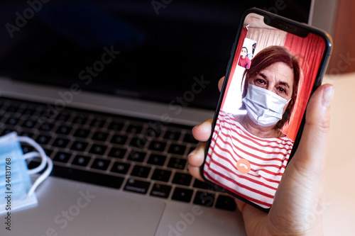 Videochiamata da cellulare tra madre con mascherina protettiva e figlia, sullo sfondo un computer con un mascherina chirurgica sopra  photo