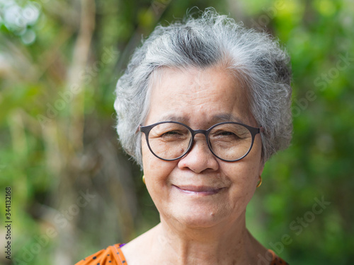 A portrait of an elderly woman wearing eyeglasses, smiling and looking at the camera while standing in a garden. Space for text. Concept of old people and healthcare