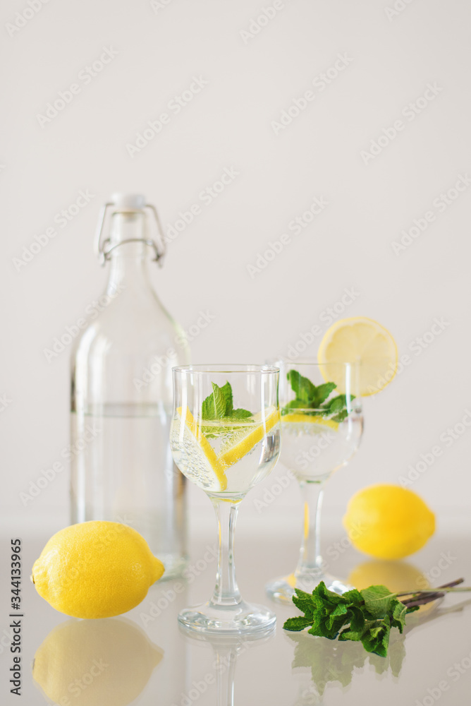 Bottle of mineral water and two glasses of sparkling drink with lemon and fresh mint on glass table. Citrus lemonade