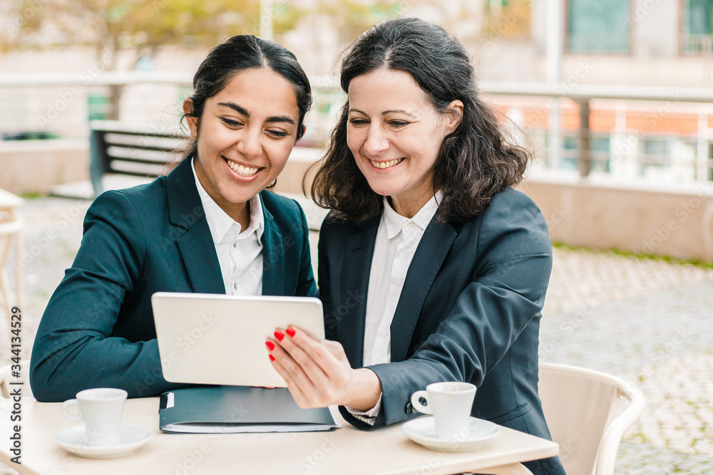 Content businesswomen with tablet pc in outdoor cafe. Cheerful female colleagues sitting at table, drinking coffee and using digital tablet in outdoor cafe. Wireless technology concept Content businesswomen with tablet pc in outdoor cafe. Cheerful female colleagues sitting at table, drinking coffee and using digital tablet in outdoor cafe. Wireless technology concept