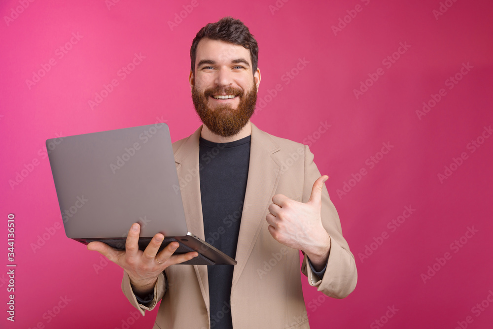 Cheerful bearded man is holding his laptop and showing like gesture on pink background. Cheerful bearded man is holding his laptop and showing like gesture on pink background.