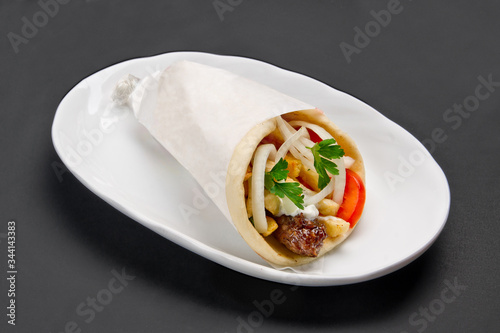 Meat, French fries and vegetables wrapped in pita with sauce on white ceramic plate on grey background