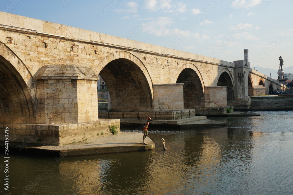 Stone Bridge (Kamen Most) spans the Vardar River, view from below. Elegant arches bridge leads to the historic Ottoman quarter of Skopje. Sculpture of Diving woman into the river in front of bridge.