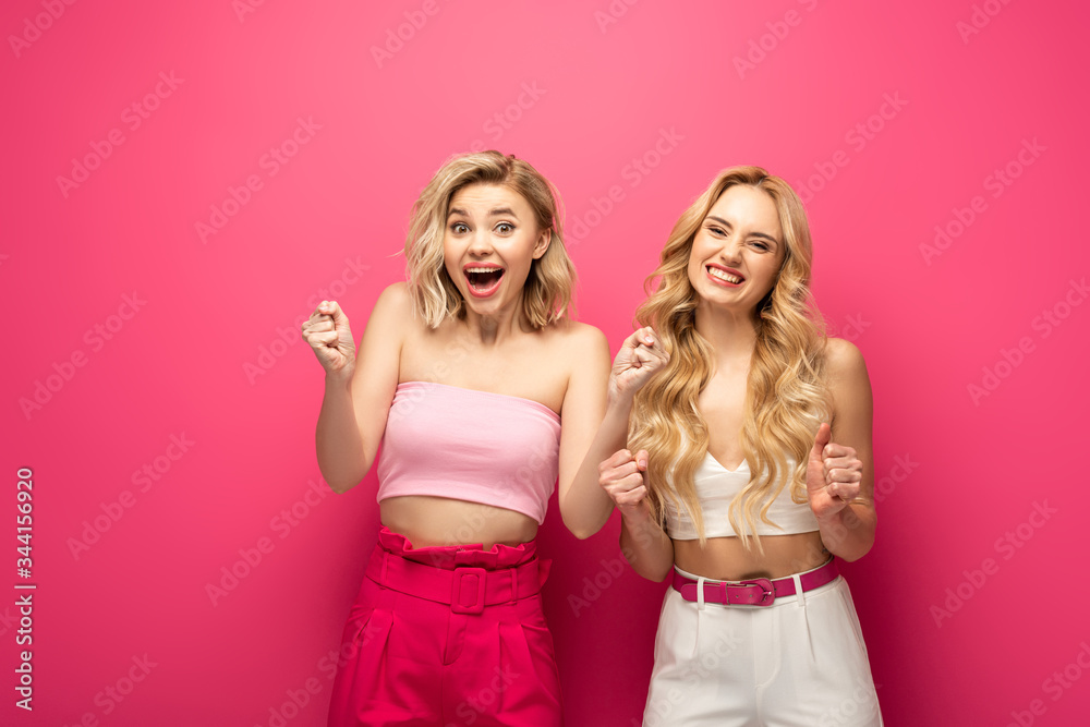 Happy blonde friends showing yeah gesture at camera on pink background Happy blonde friends showing yeah gesture at camera on pink background