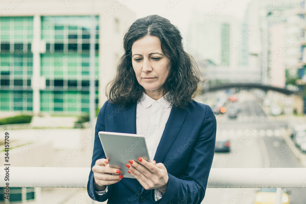 Focused woman using tablet pc. Serious middle aged businesswoman using digital tablet while standing on urban city street. Wireless technology concept