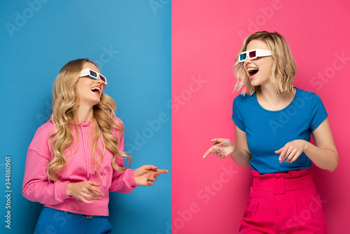 Laughing blonde sisters in 3d glasses pointing with fingers on blue and pink background
