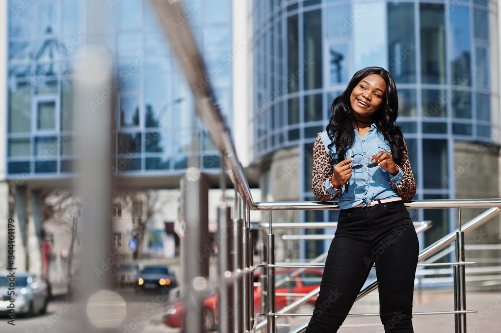 Hipster african american girl wearing jeans shirt with leopard sleeves posing at street against modern office building with blue windows.
