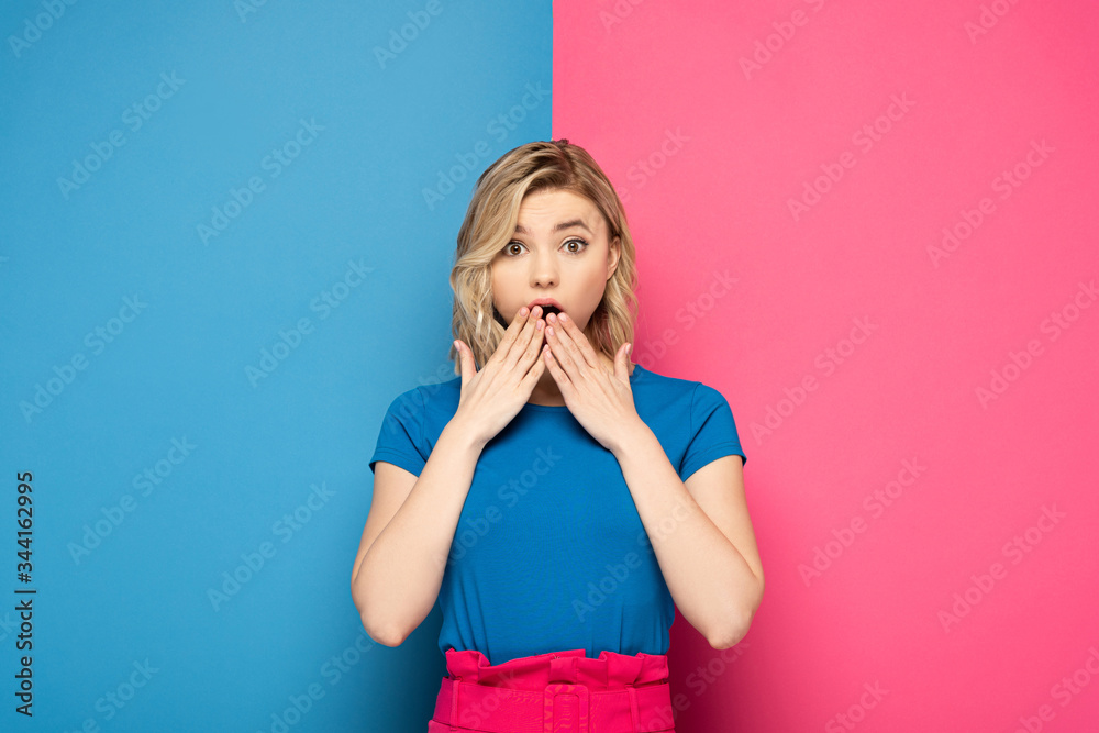 Surprised blonde woman looking at camera on pink and blue background Surprised blonde woman looking at camera on pink and blue background