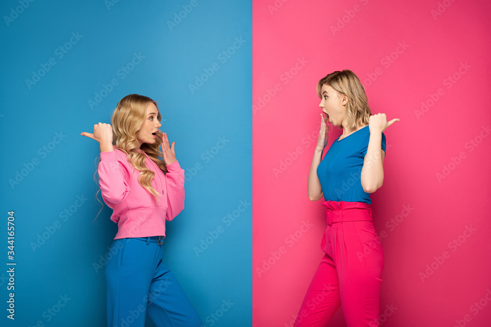 Shocked blonde girls pointing with fingers on pink and blue background Shocked blonde girls pointing with fingers on pink and blue background