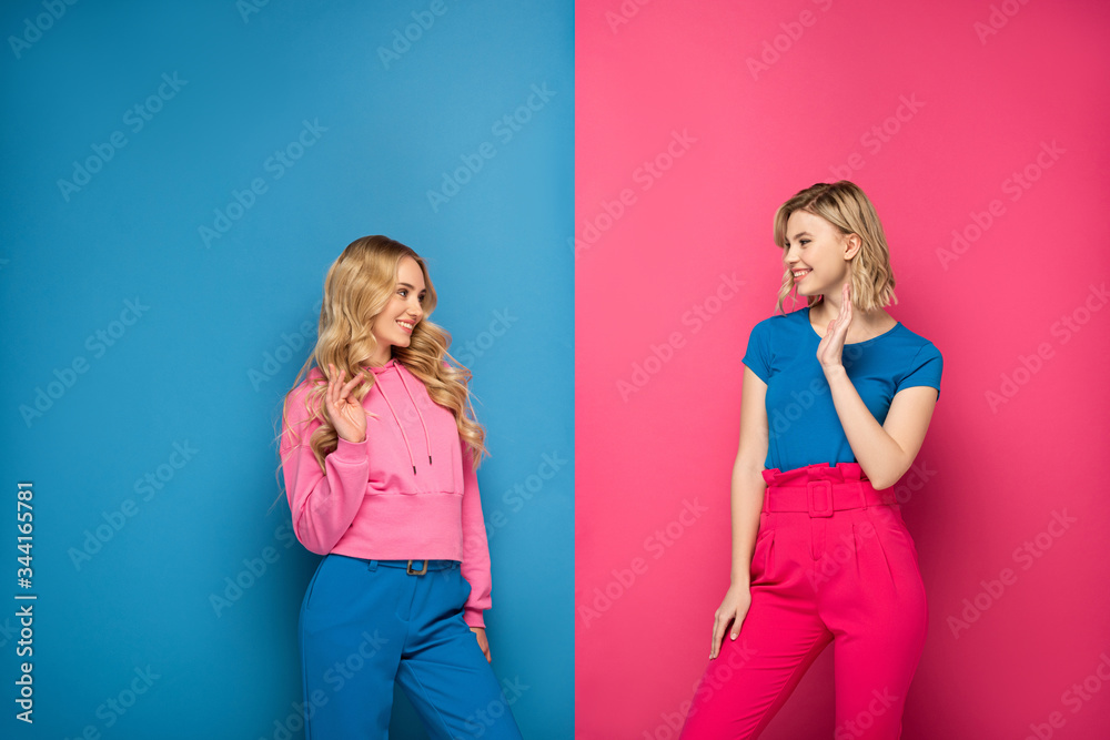 Smiling blonde girls waving hands at each other on pink and blue background Smiling blonde girls waving hands at each other on pink and blue background