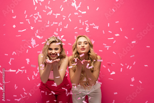 Smiling blonde girls blowing air kiss at camera under falling confetti on pink background
