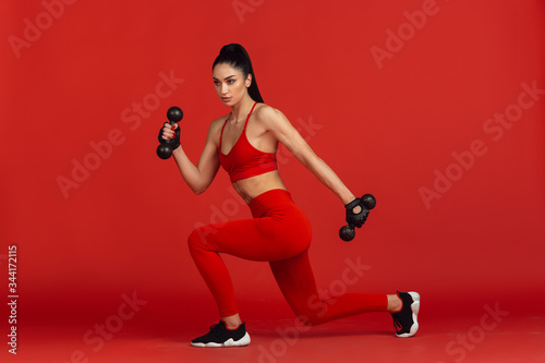 Graceful. Beautiful young female athlete practicing in studio, monochrome red portrait. Sportive fit brunette model with weights. Body building, healthy lifestyle, beauty and action concept.