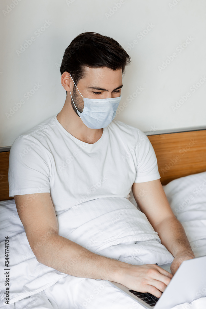 Freelancer in medical mask working with laptop in bedroom