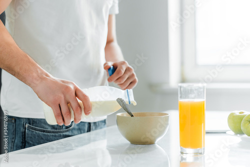 Cropped view of man pouring milk in bowl near orange juice and apples on table