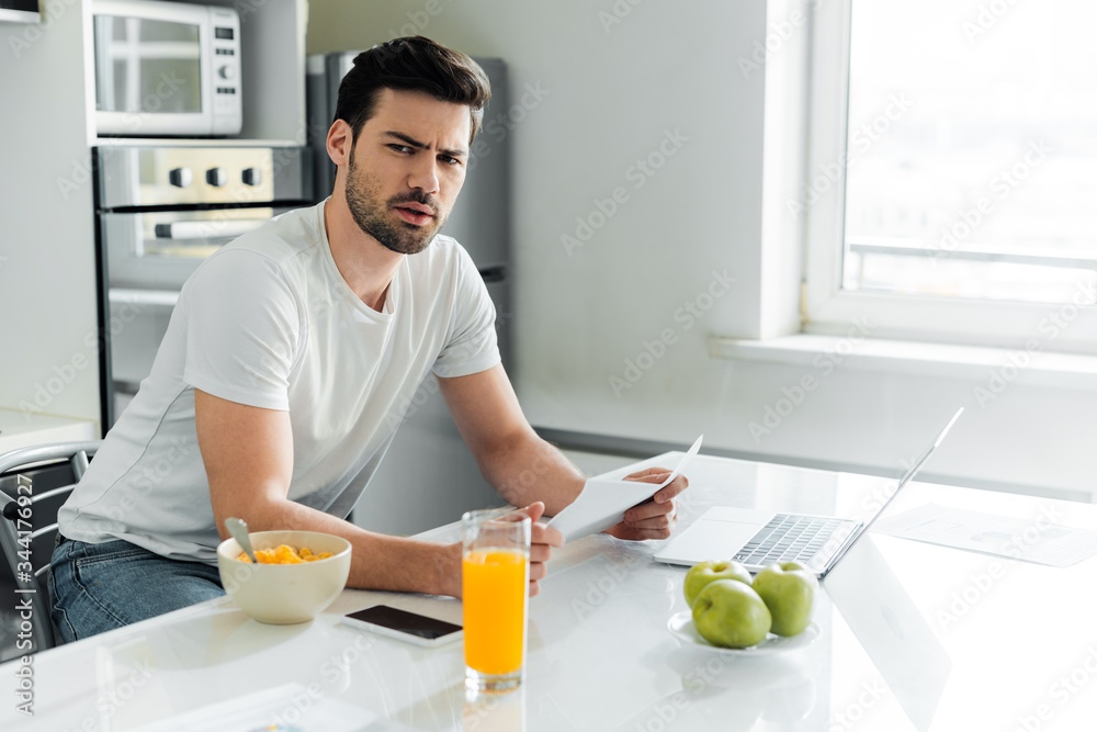 Pensive freelancer looking at camera while holding paper near gadgets and cereals on kitchen table Pensive freelancer looking at camera while holding paper near gadgets and cereals on kitchen table