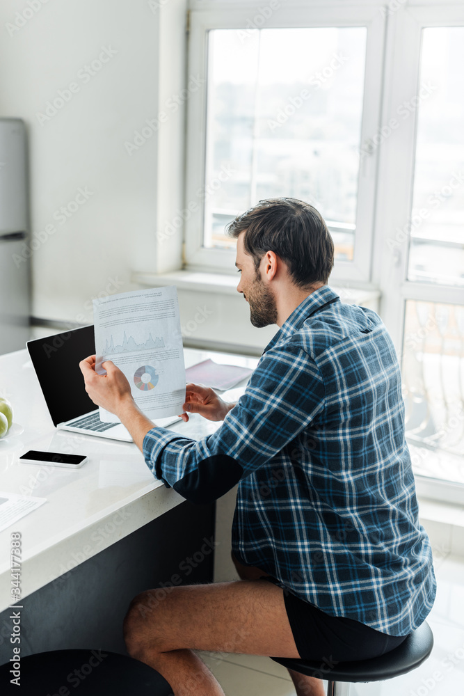 Side view of freelancer in plaited shirt and panties holding paper with charts near laptop and smartphone on table in kitchen Side view of freelancer in plaited shirt and panties holding paper with charts near laptop and smartphone on table in kitchen