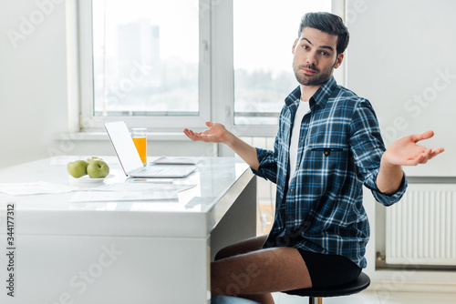 Side view of freelancer in shirt and panties showing confused sign at camera near laptop and papers on table