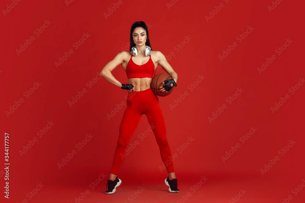 Beautiful young female athlete practicing in studio, monochrome red portrait. Sportive fit brunette model with ball and headphones. Body building, healthy lifestyle, beauty and action concept.