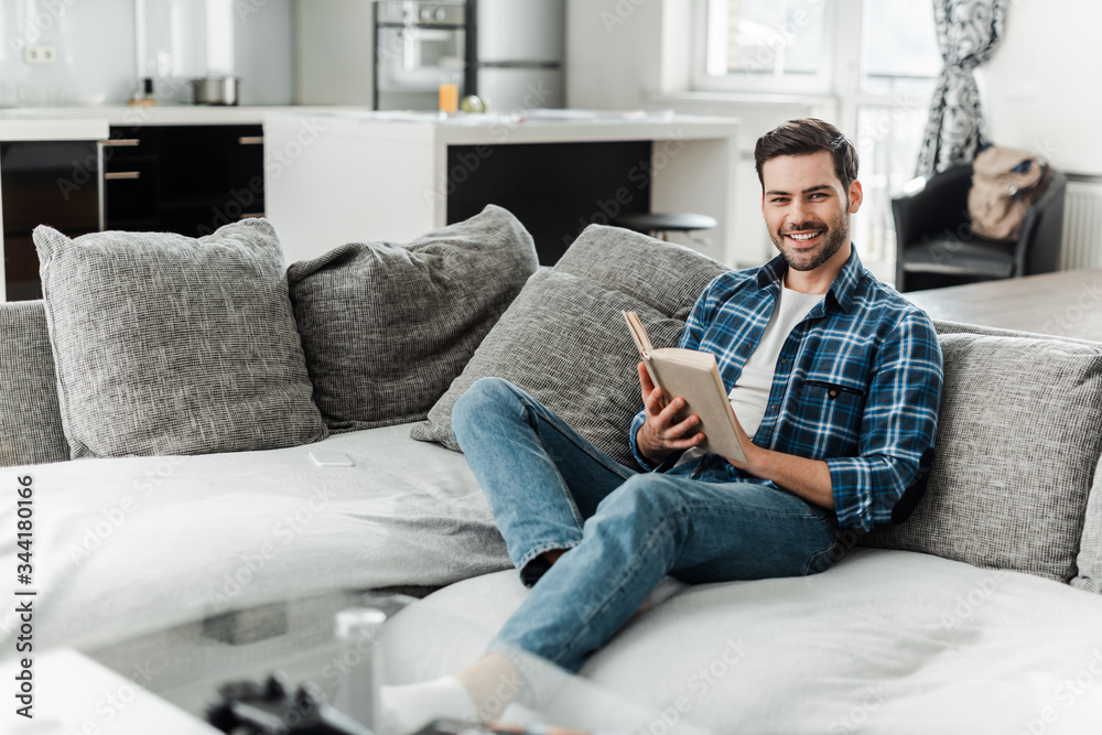 Selective focus of handsome man smiling at camera while reading book on couch at home
