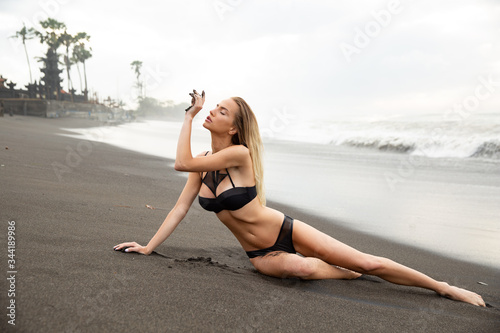 Beautiful sexy girl model lies on black sand beach, in a black swimsuit, with a tanned body, long blonde hair, magnificent breasts and puffy lips. Healthy nature, ocean waves, clouds on Bali island. © evgennish