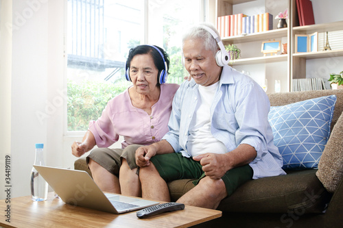 Elderly couple Wearing headphones to watch media online on a laptop In the living room at home Both are happy in life after retirement. Concept of health insurance, social security