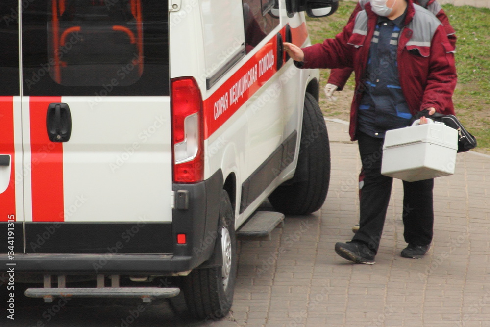 Fototapeta premium A doctor in red outfit with a white suitcase and medical mask open ambulance car with Russian inscription AMBULANCE, people antiviral prevention