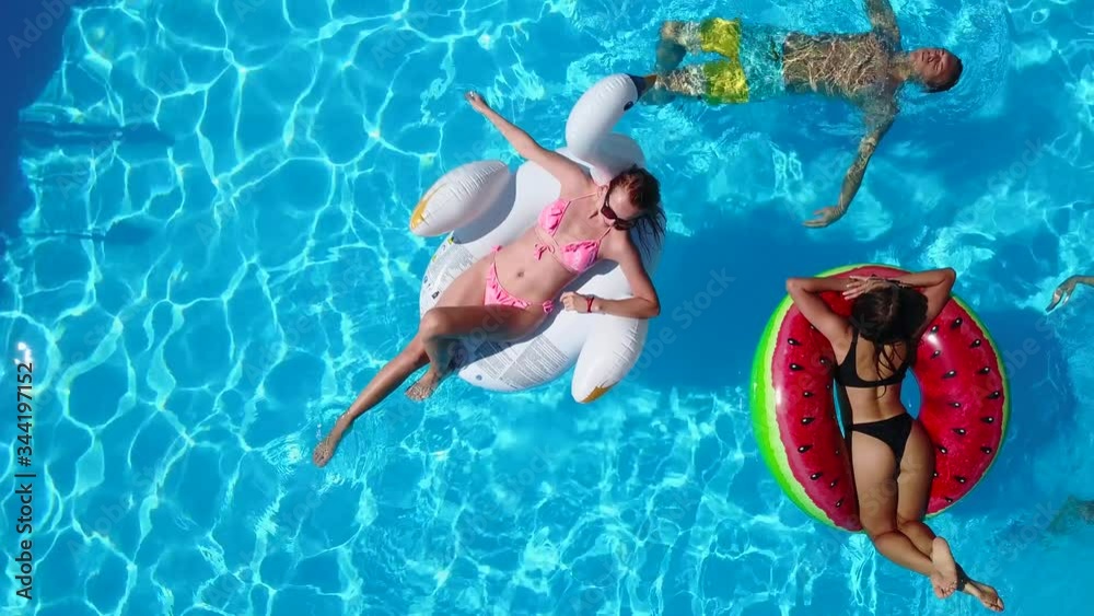 Aerial. Friends chilling in swimming pool with inflatable flamingo, swan, mattress. Happy young people bathe on floating mattresses in luxury resort. View from above. Girls in bikini sunbathing in sun