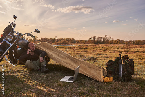 A traveler on a motorcycle talking on the phone working on a laptop admires the sunset