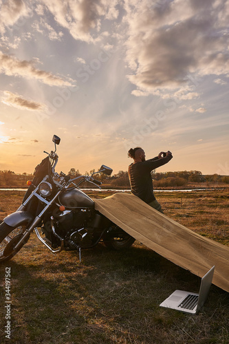 A traveler on a motorcycle talking on the phone working on a laptop admires the sunset