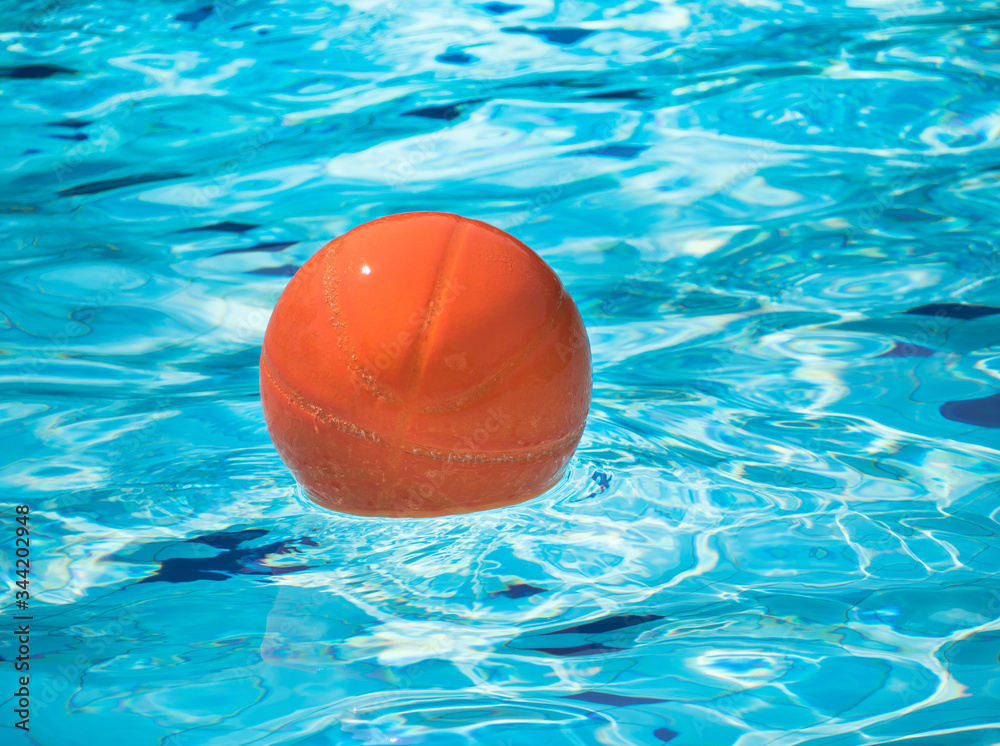 Orange beach ball floating in blue swimming pool. Ball in pool on sunny day, Summer holiday vacation.