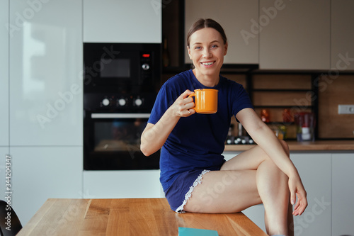 Positive young woman enjoying a cup of tea while sitting on the table in modern kitchen staying home photo