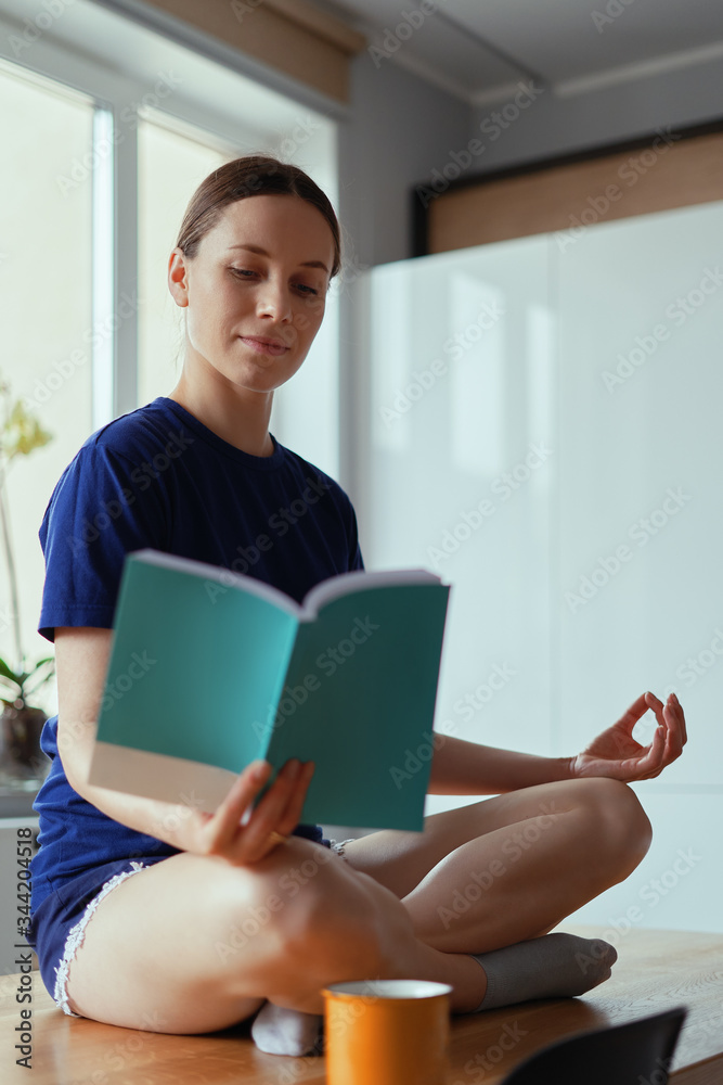 Fototapeta premium Young woman relaxing in yoga poses reading , sitting on a wood table in a modern interior with windows in the background