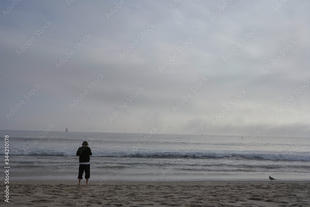 Tourist on the beach in Santa Monica in California, standing barefoot in the sand photographing the foaming sea and the beautiful horizon with clouds and seagulls and a boat in the pacific