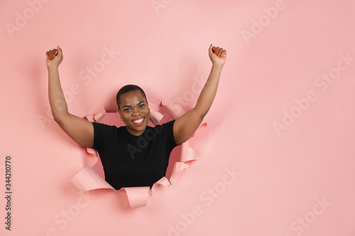Winner. Cheerful african-american young woman poses in torn coral paper background, emotional and expressive. Breaking on, breakthrought. Concept of human emotions, facial expression, sales, ad. © master1305