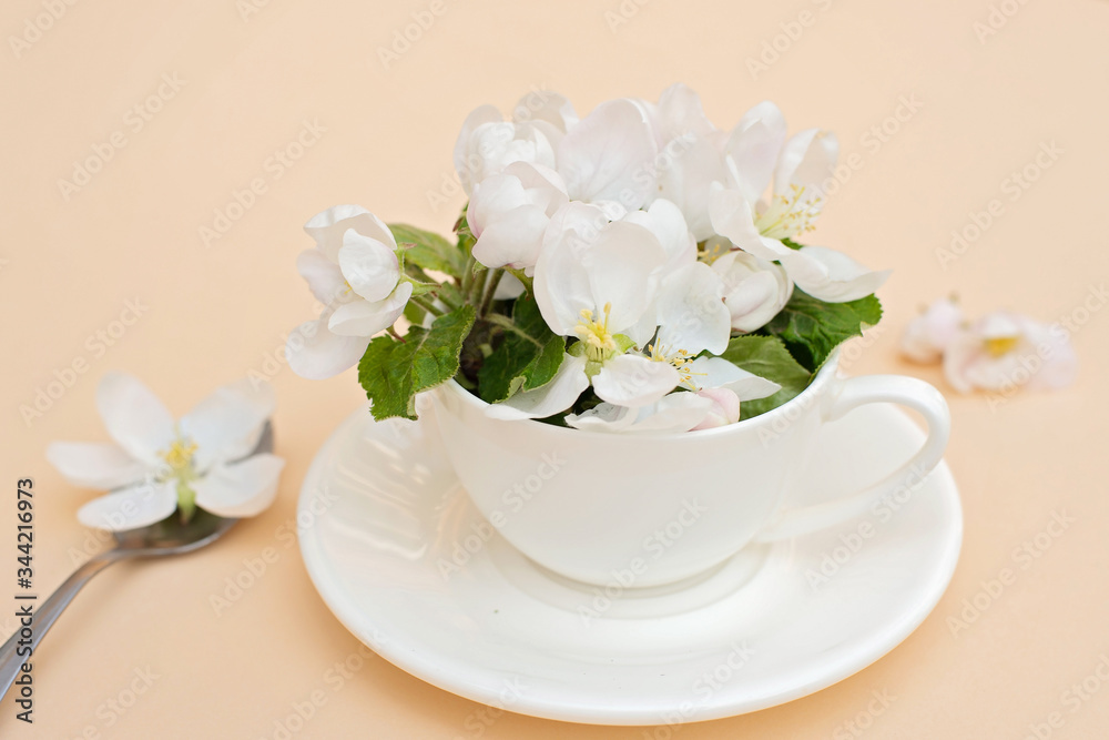 White spring apple blossoms blooming flowers in a coffee cup with a spoon on a beige background. Spring summer concept. Greeting card.