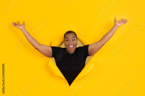 Happy greeting. Cheerful african-american young woman poses in torn yellow paper background, emotional expressive. Breaking on, breakthrought. Concept of human emotions, facial expression, sales, ad. © master1305