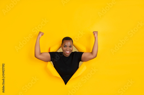 Strong. Cheerful african-american young woman poses in torn yellow paper background, emotional and expressive. Breaking on, breakthrought. Concept of human emotions, facial expression, sales, ad. © master1305