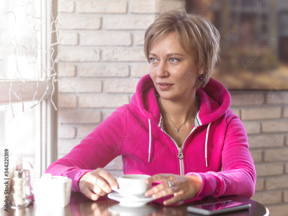 A young woman in a purple hoodie sits in a cafe and drinks coffee from a white cup. Daytime, lunch, portrait, close-up, soft light. Healthy lifestyle and sports concept. A young woman in a purple hoodie sits in a cafe and drinks coffee from a white cup. Daytime, lunch, portrait, close-up, soft light. Healthy lifestyle and sports concept.