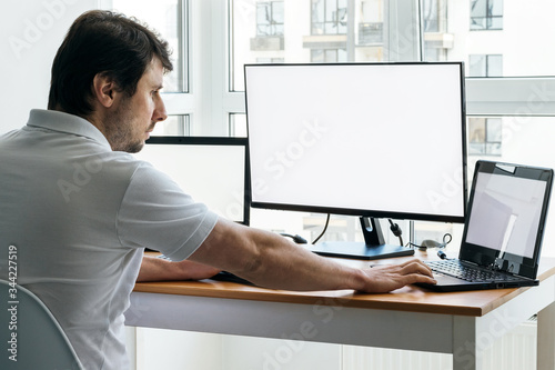 A man in a white t-shirt is sitting at a workplace with two laptops and a monitor near the window. Remote work from home in quarantine