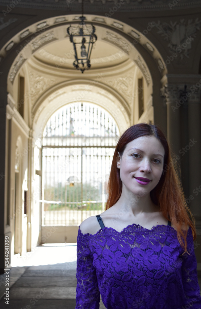 Portrait of a beautiful young woman with long red hair and a purple lace dress with an Italian stone and marble neoclassical palace entrance in the background.