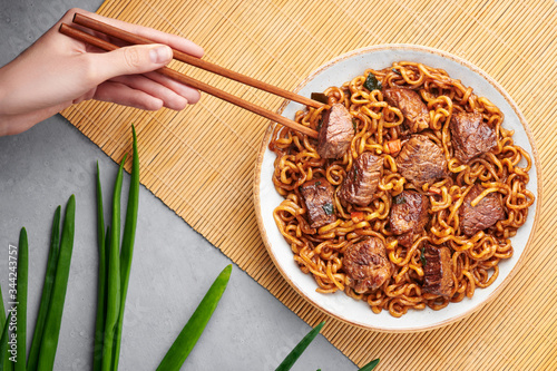 Ram-Don or Chapaguri noodles with beef steak in white bowl on grey concrete backdrop. Jjapaguri is a popular south korean dish with ramen and udon noodles and beef steak. Female hand with chopsticks