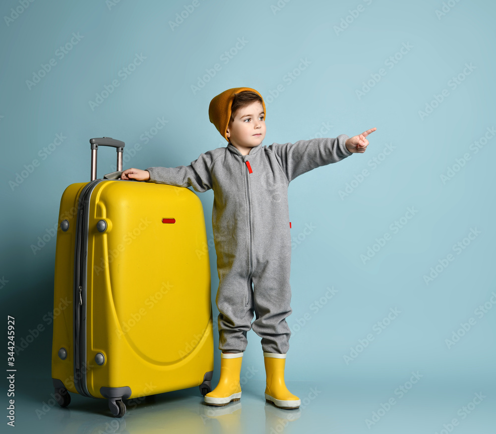 Little kid in gray overall, yellow rubber boots and hat. Holding big suitcase wheeler pointing at copy space Little kid in gray overall, yellow rubber boots and hat. Holding big suitcase wheeler pointing at copy space