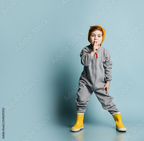 Frolic kid brunet boy in orange hat, gray overall and yellow rubber boots gestures shows Shh sign over blue background